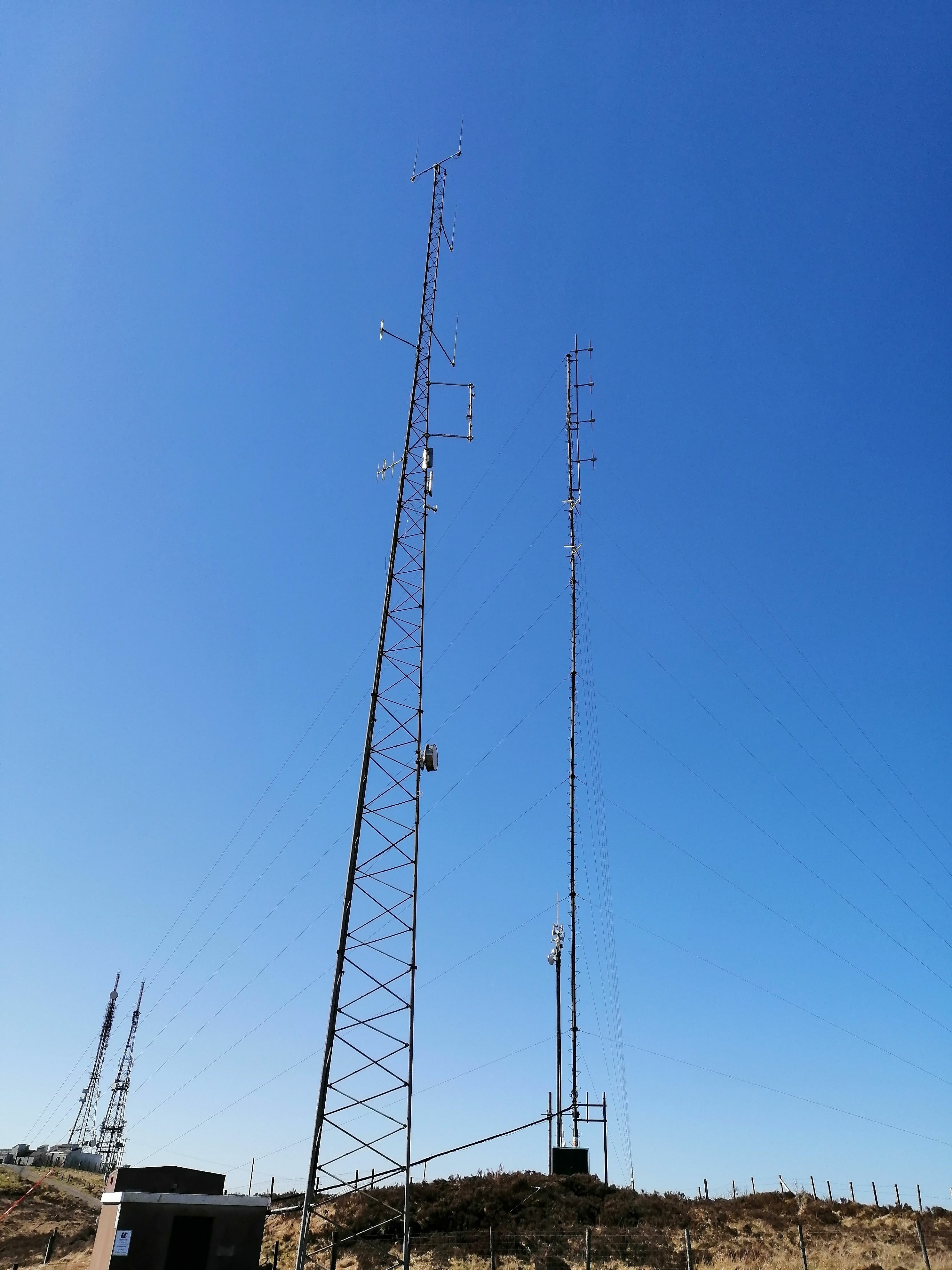 Q Radio has a taller stayed mast, whilst the Ulstercom mast is a triangular cross-sectioned free-standing tower.