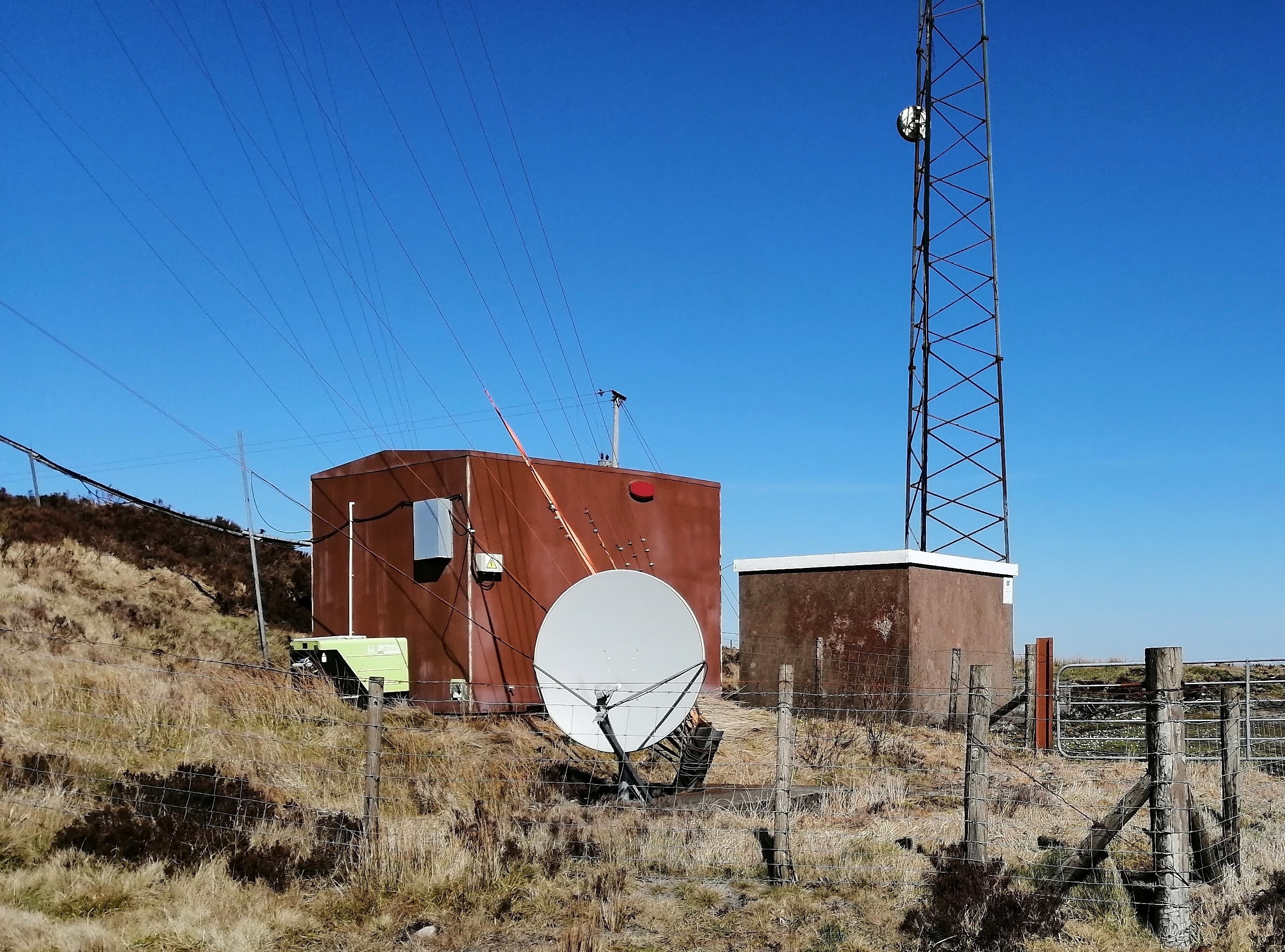 Q Radio has a newer 'pre-fab' type building, with a satellite dish and small generator outside. Cables run up one of the mast stays from the building. Ulstercom have an older concrete building with cables running up the side to their mast. A sign on the side offers contact information for them.