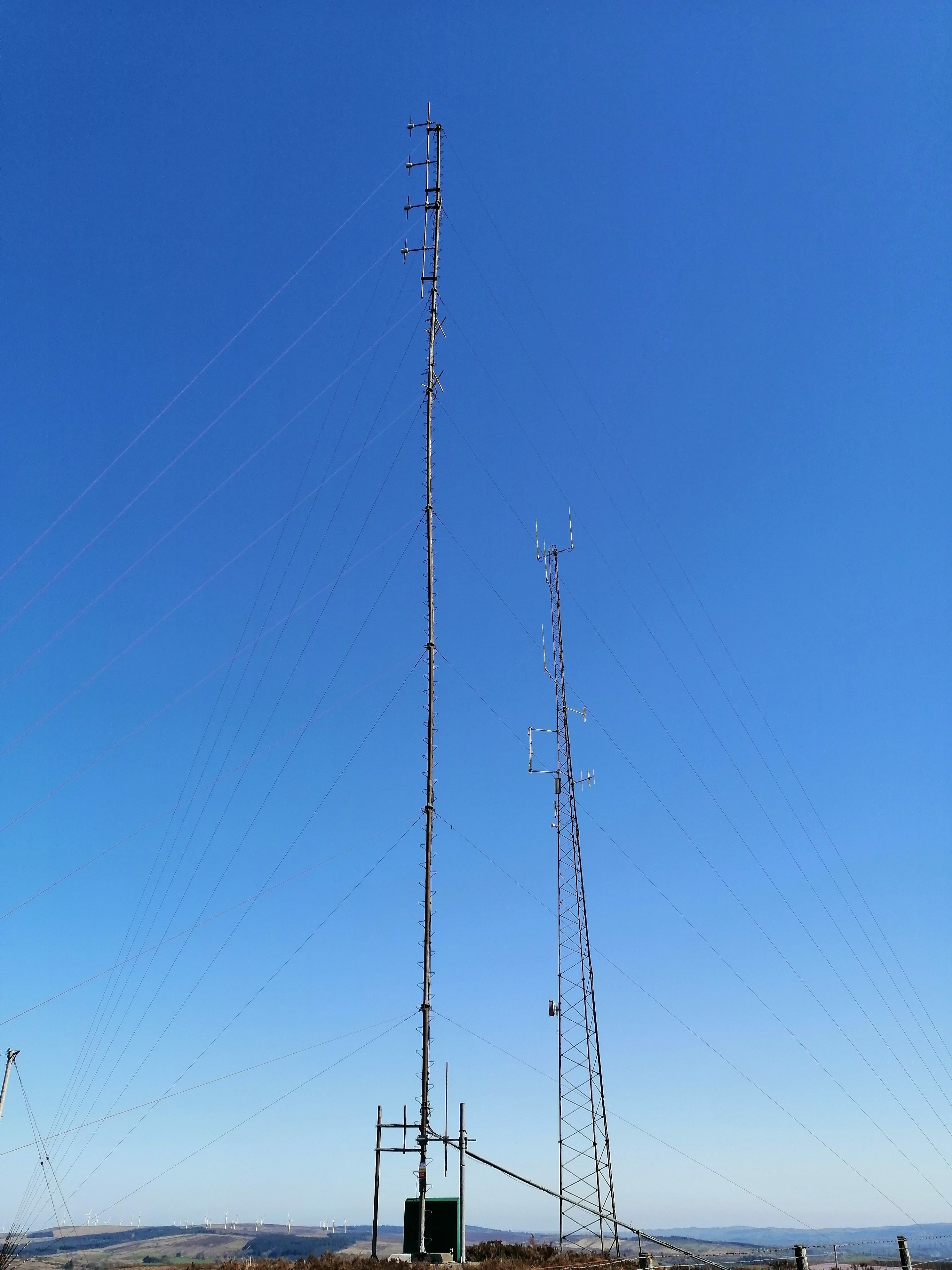 Wide view of the masts. The Q Radio mast is fixed the the ground by two additional smaller poles at it's base. A wind farm is visible on the distant hill.