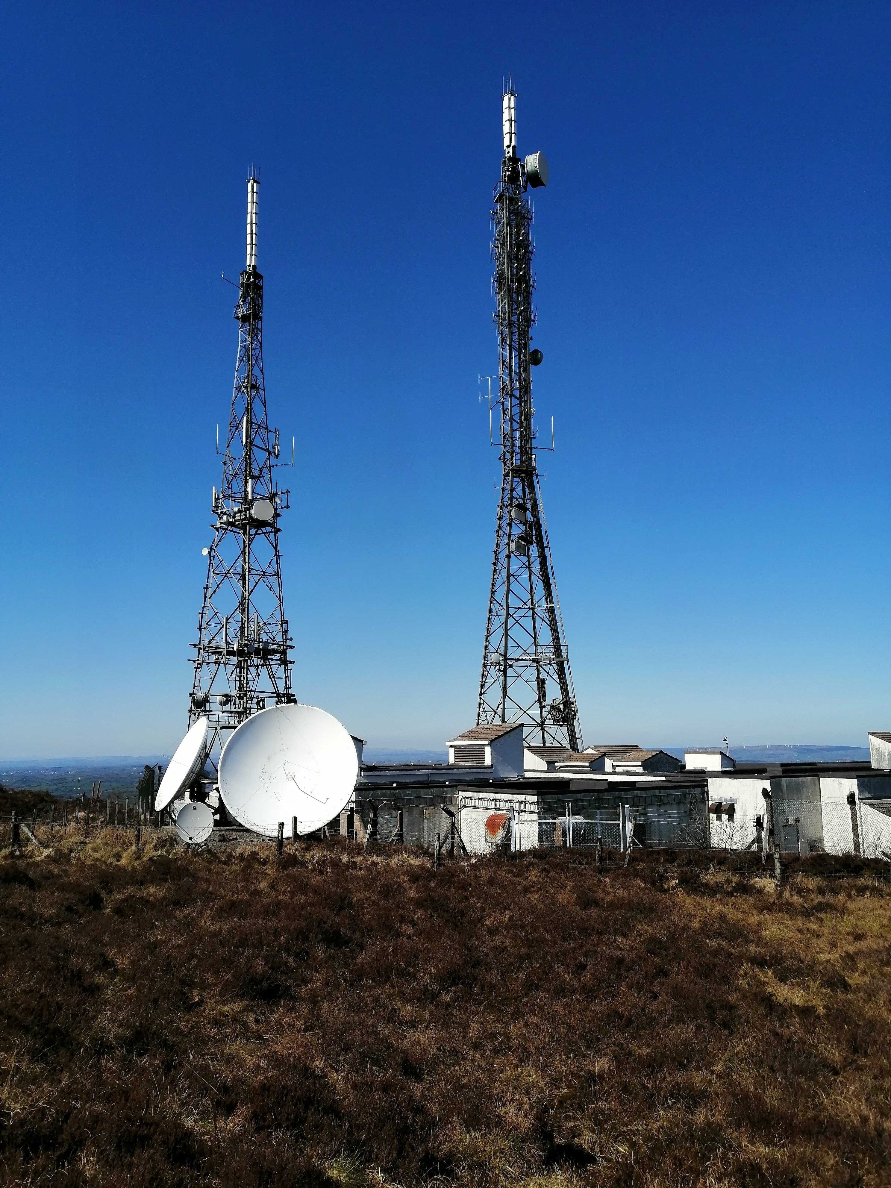 Two large, free-standing towers, 3 satellite dishes and a sprawling concrete building form surrounded by a tall chain-link fence make up the Arqiva conpound. The building has some odd obtrusions on the roof which I can only assume are for ventilation purposes. Another distant wind farm is visible in the distance.