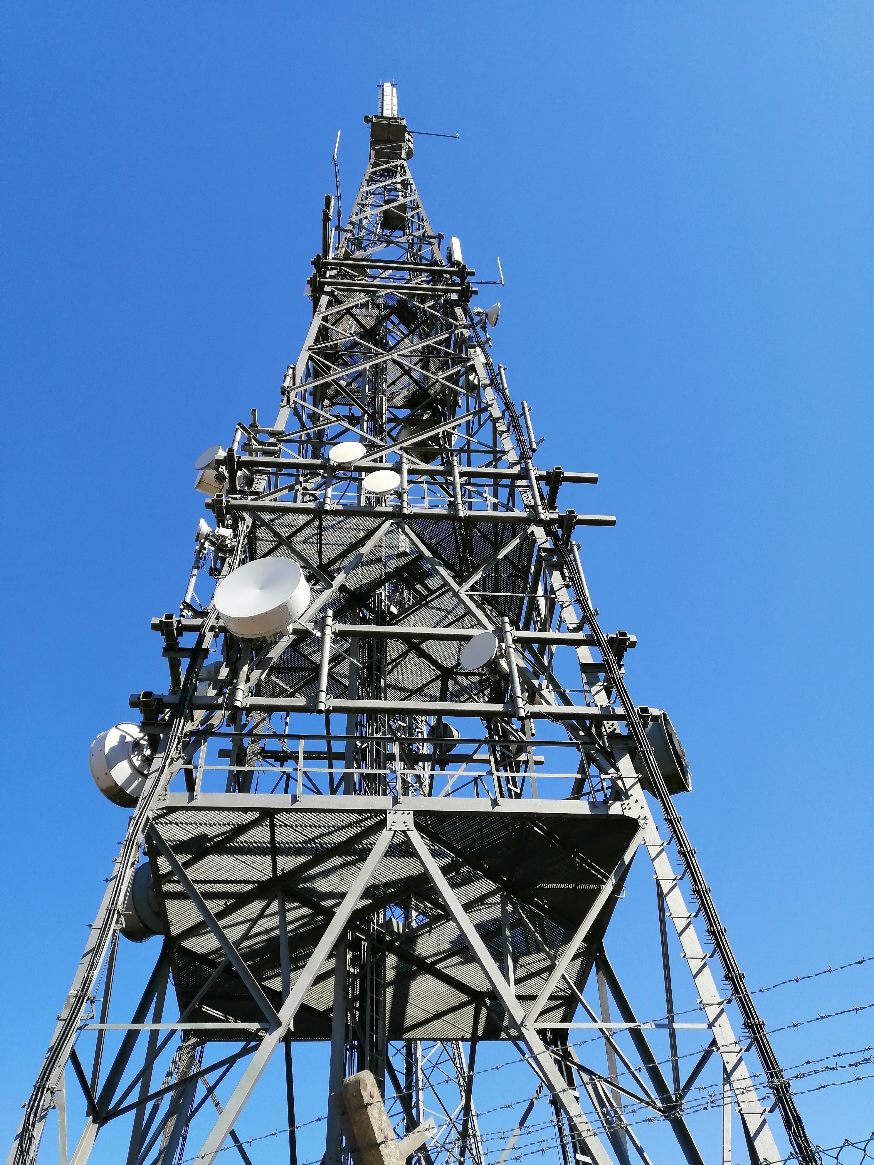 Looking up Mast B. Some vertical mobile cell antennas are visible, as well as microwave dishes of various sizes. Some of them appear to be damaged. There's a large group of panel antennas at the very top for DTT.