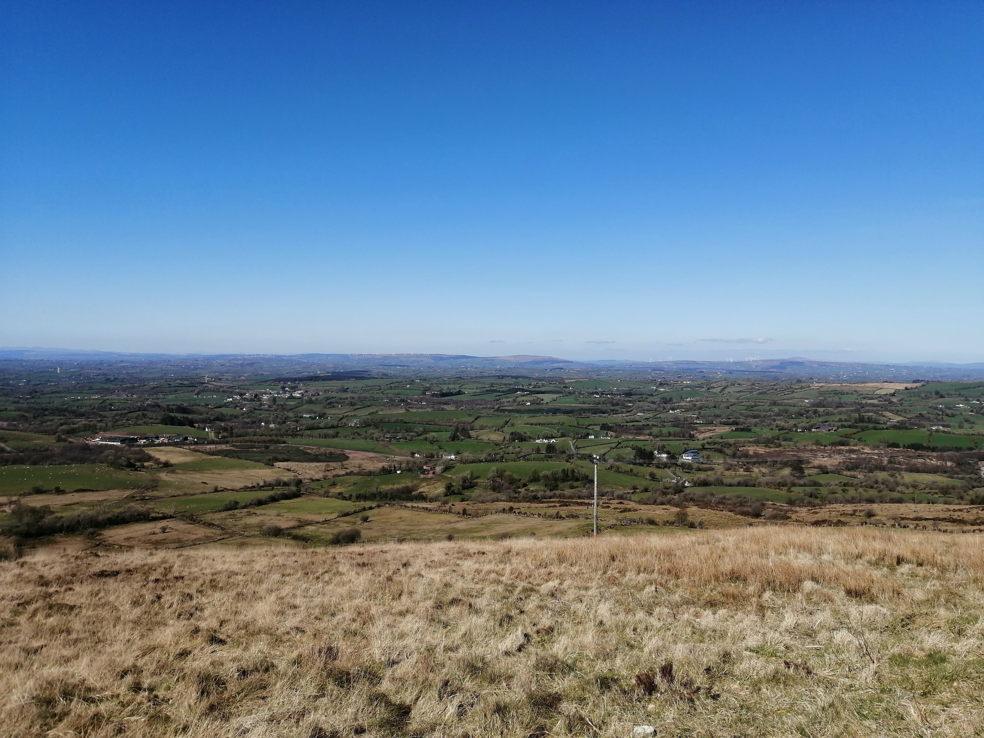View from the peak. This consists of green fields with distant houses, giving way to mountains (with yet another wind farm) and a perfect blue sky. Some white clouds are beginning to appear on the horizon.