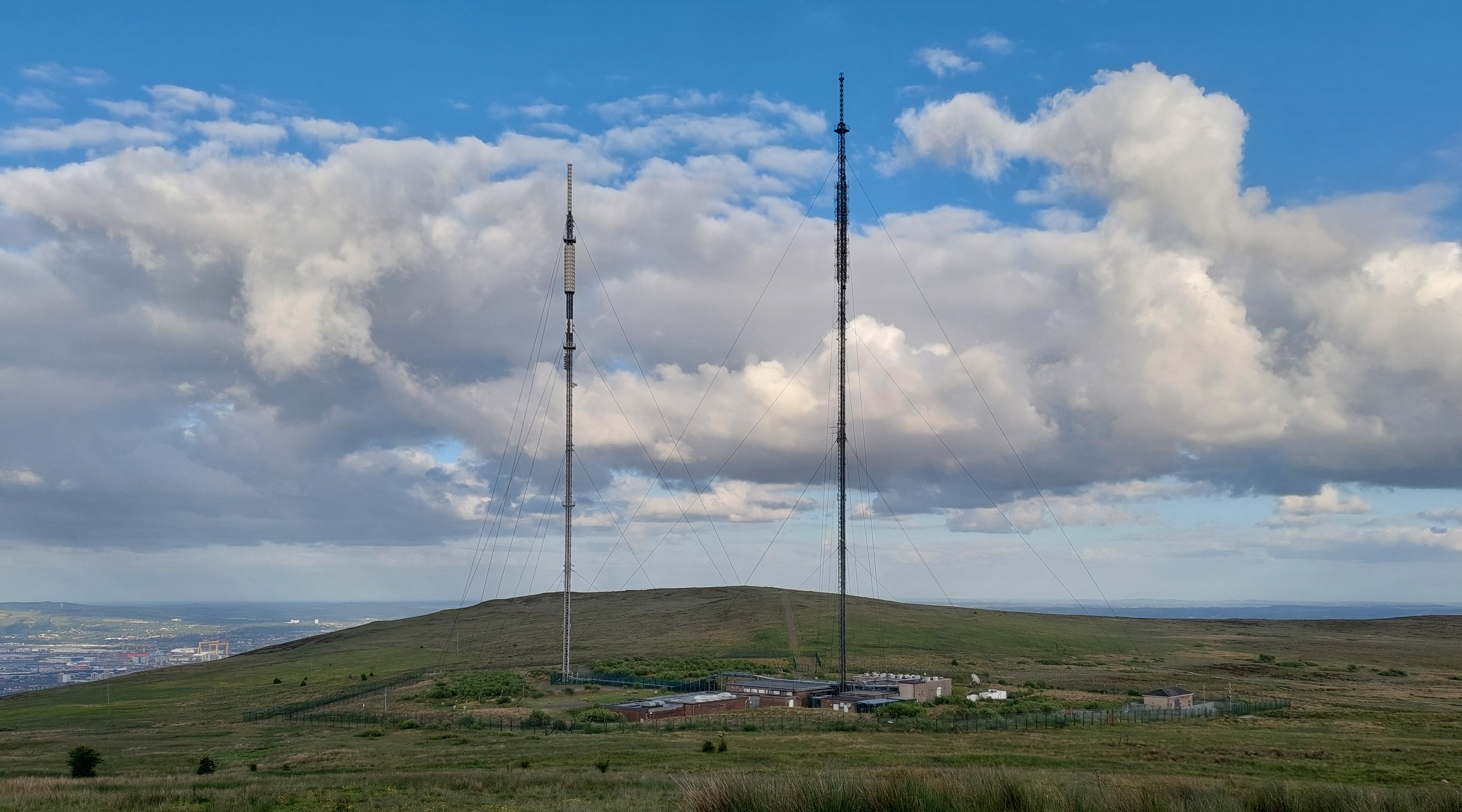 Similar to previous - stayed masts surrounded by buildings and fencing. Black Mountain peak visible behind - as well as the famous Harland & Wolff Cranes.