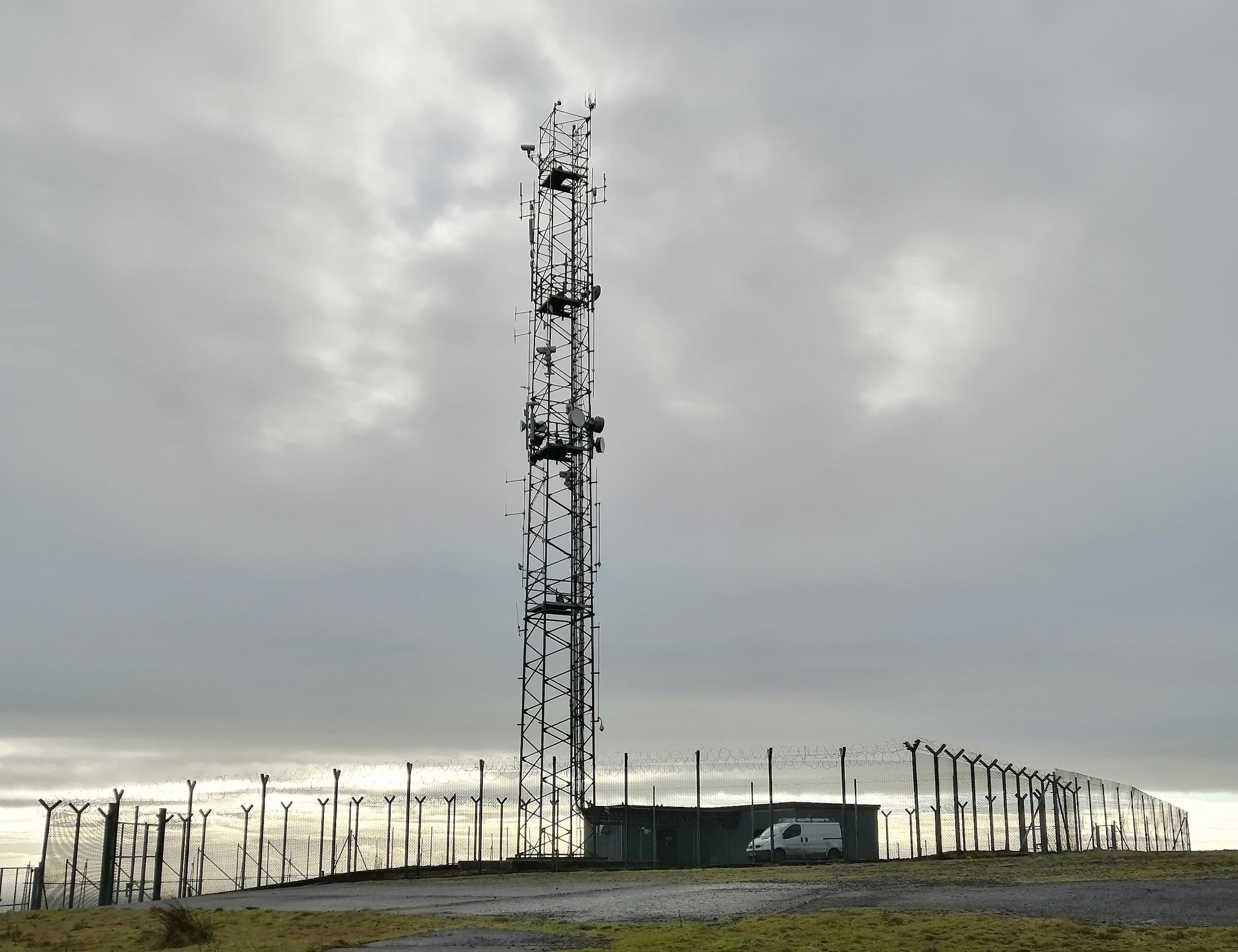 Single self-supporting mast, with small transmitter building surrounded by chain-link fence. A van can be seen in the yard.